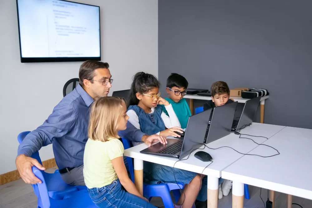 Professor sentado em uma cadeira, mexendo no computador com outros 4 alunos